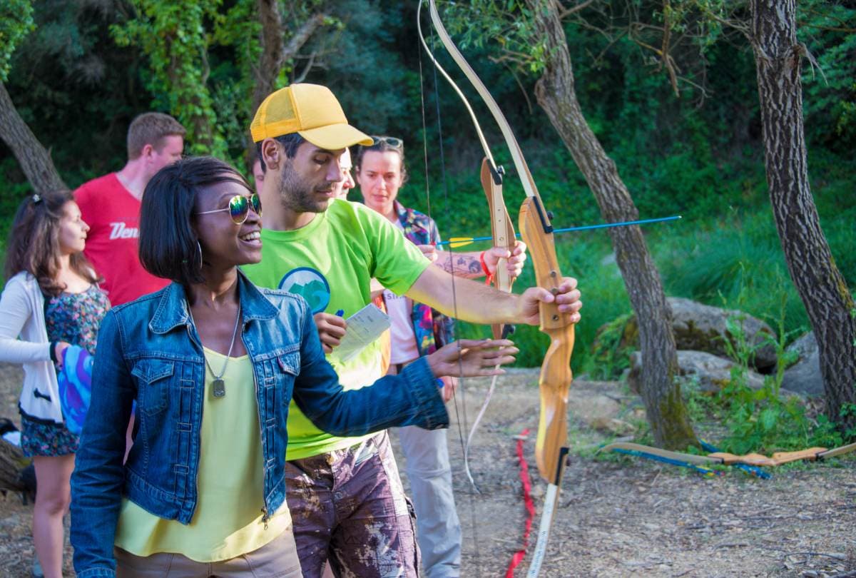 Instructor in a green tee helping a woman aim a wooden bow during an outdoor group archery lesson