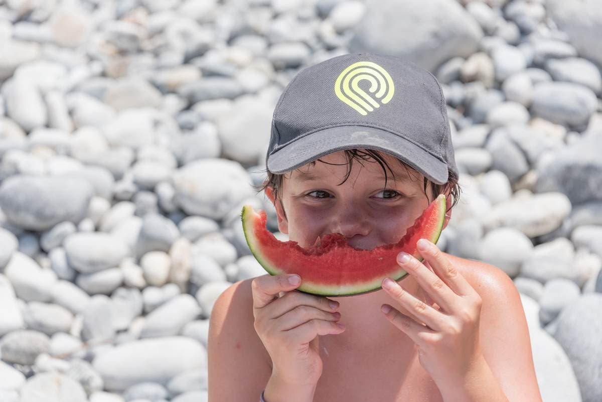 Jongen met een Explore Messinia-pet die een plak watermeloen eet op een kiezelstrand bij Navarino