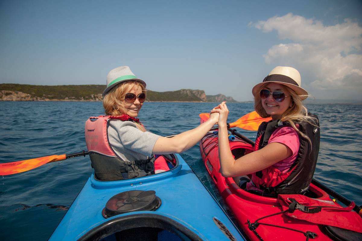 Twee vrouwen met zonnehoeden die glimlachen terwijl ze elkaars handen vasthouden tussen hun kajaks
