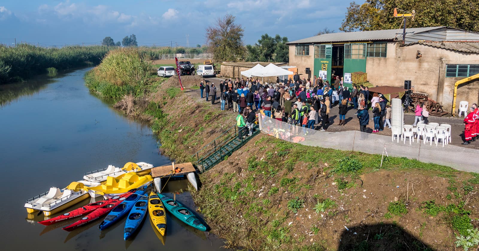 Crowd gathered for an environmental event by the Aris river, with kayaks and pedal boats moored below