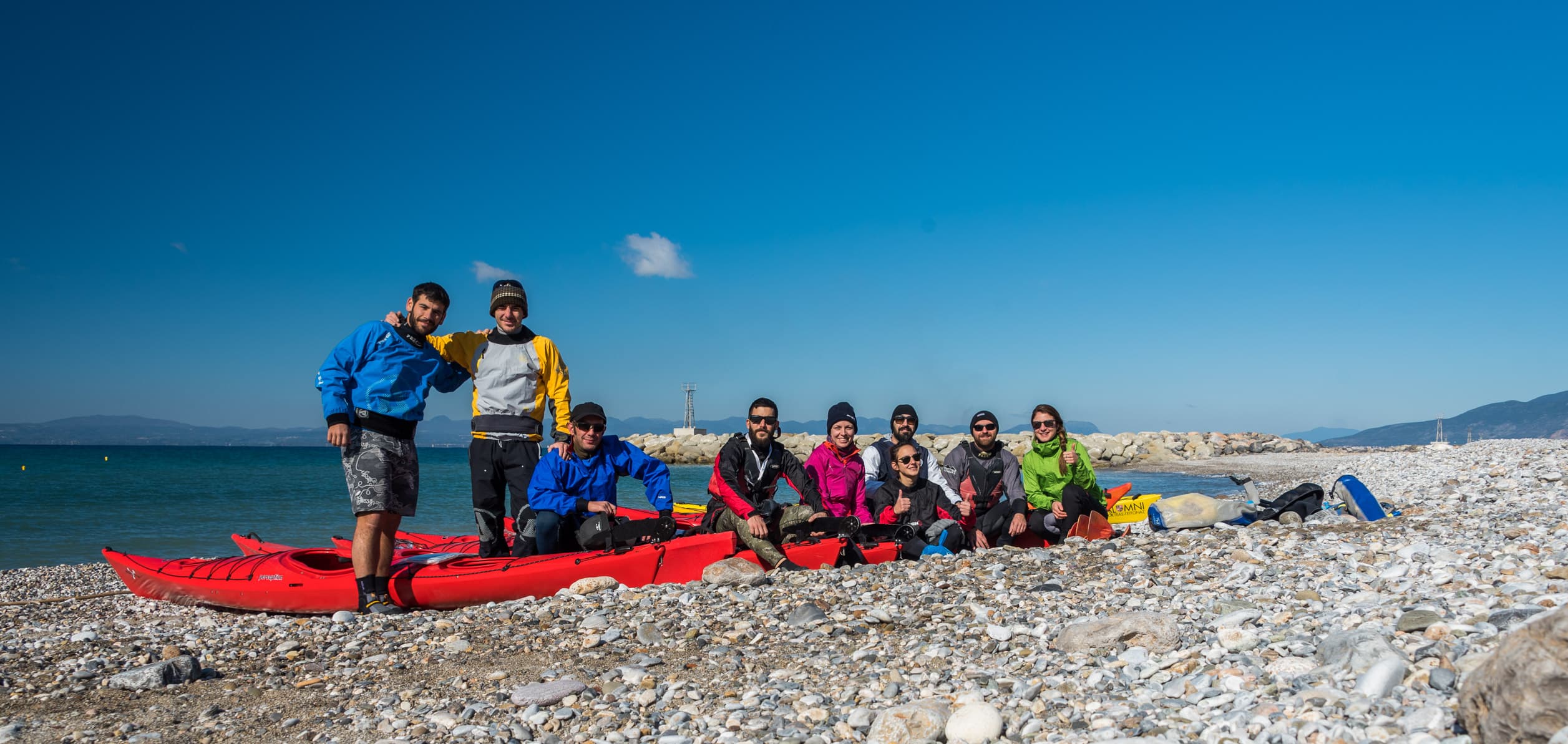 Group of sea kayakers posing on a pebble beach beside red kayaks under a clear blue sky