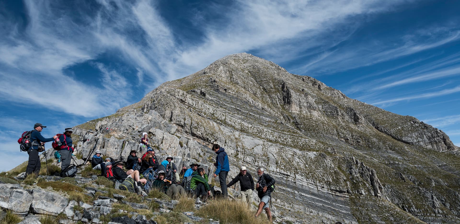 Hikers resting on a rocky ridge below the bare limestone summit of Taygetos under a bright blue sky