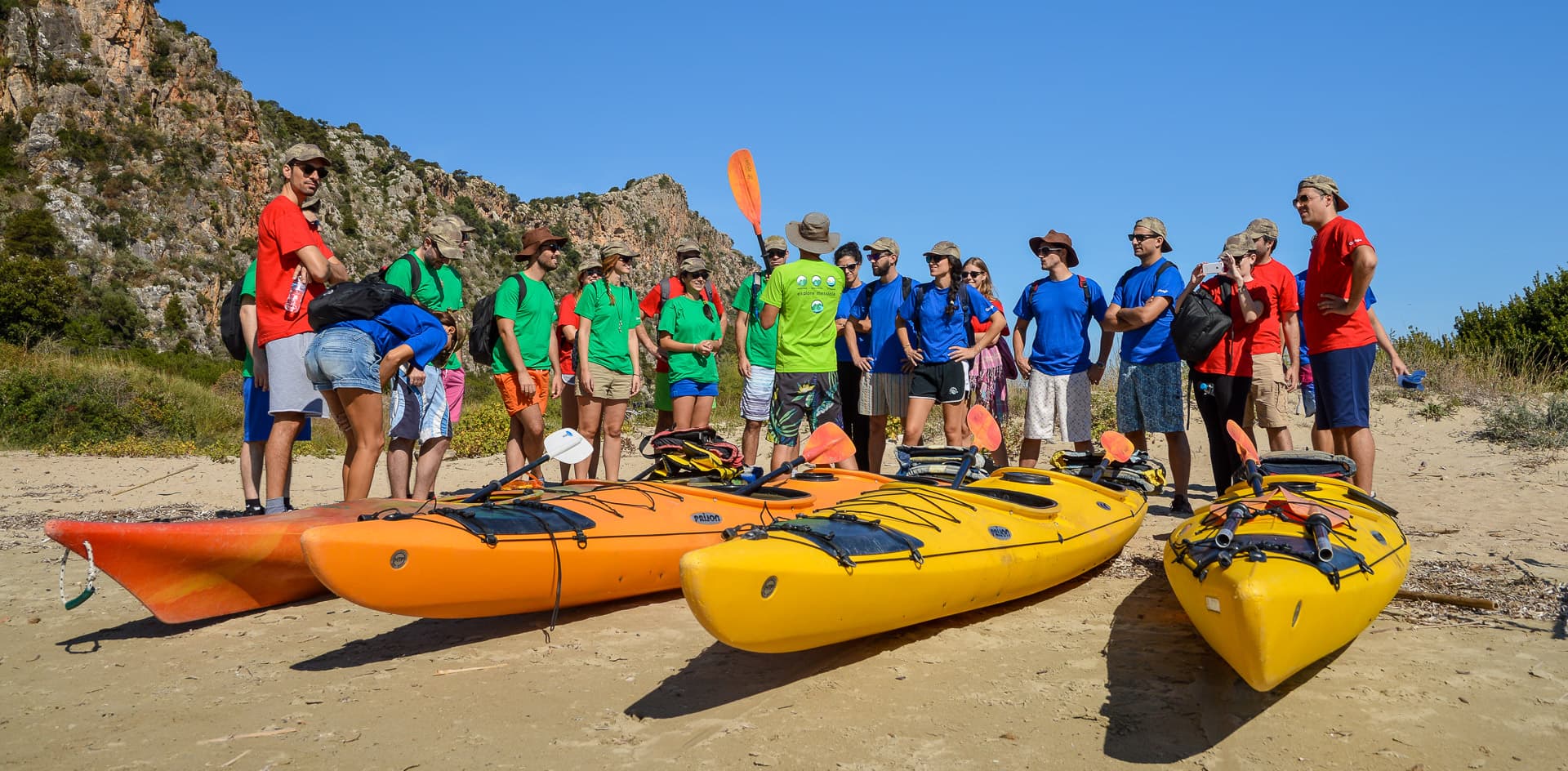 Group of participants in colored team shirts gathered around orange and yellow sea kayaks on a beach