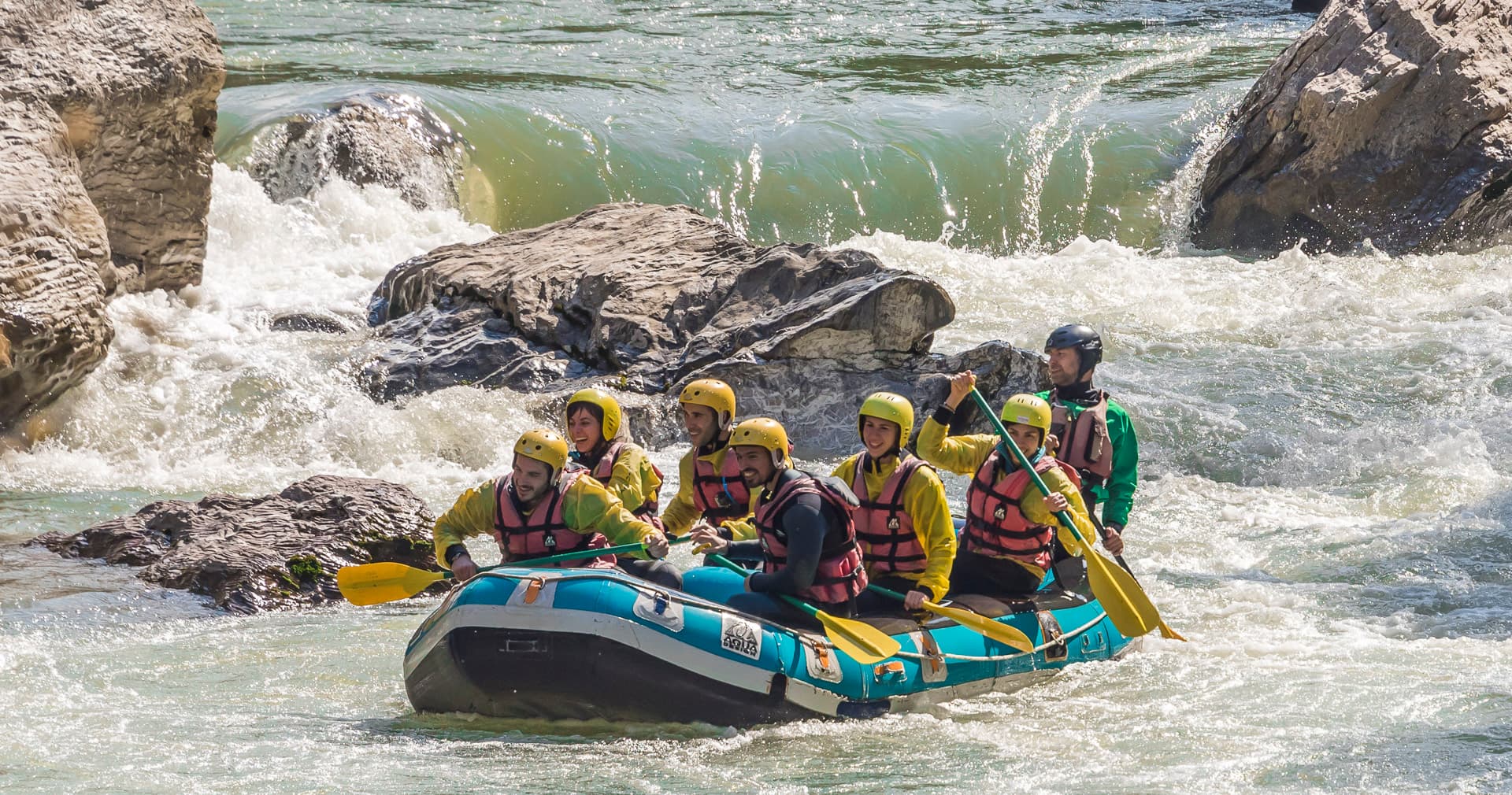 Helmeted rafting team paddling a blue raft through whitewater rapids past rocks on the Lousios river