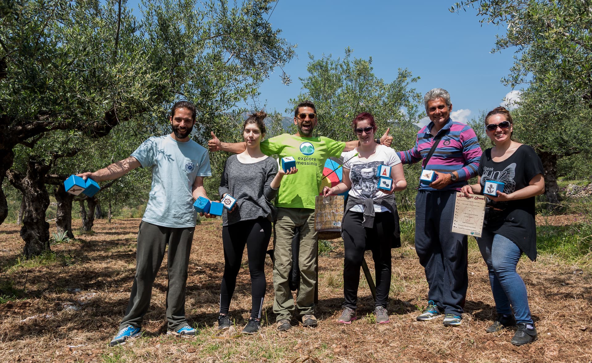 Six team-building participants holding small boxes and giving thumbs up in an olive grove near Kardamyli