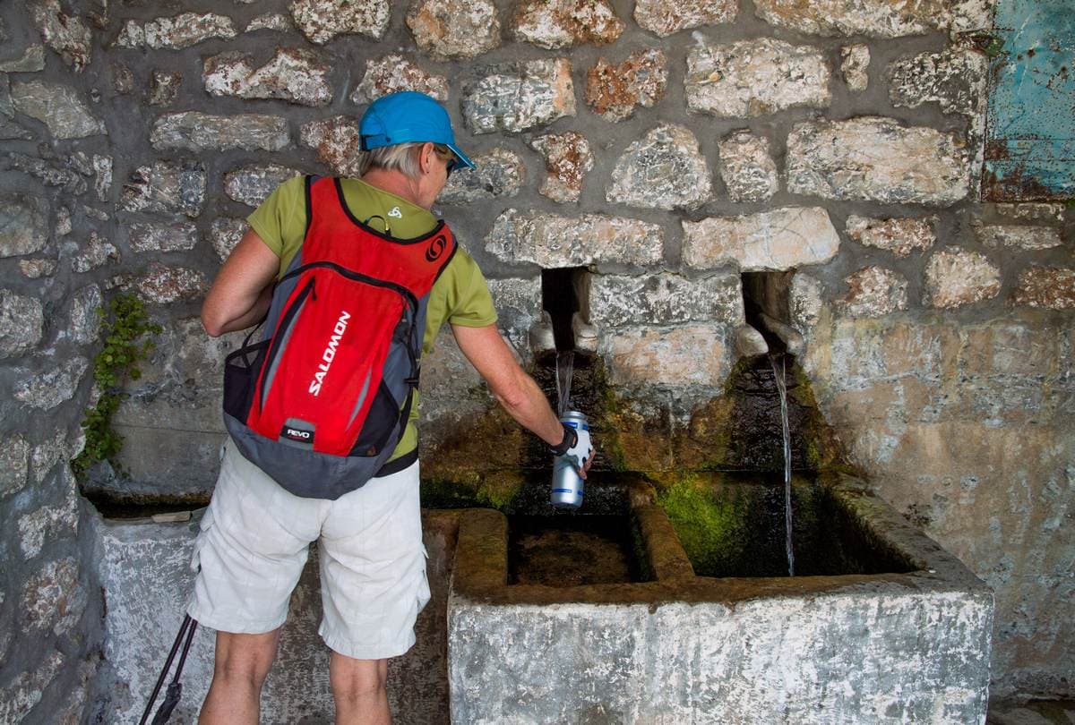 Hiker fills a water bottle at an old stone fountain with twin spouts above Kalamata