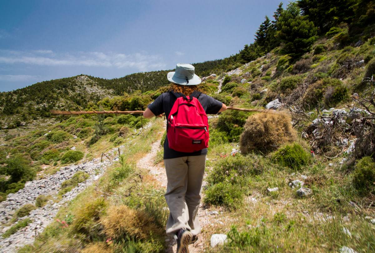 Hiker with red backpack and sun hat walks a narrow trail across green mountain slopes above Kalamata