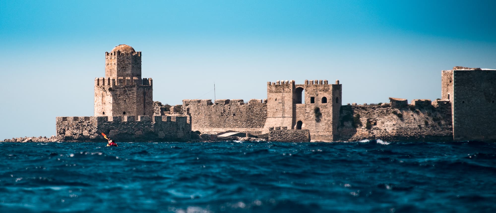 Kayaker paddling past the sea-washed stone walls and towers of Methoni Castle under a clear sky