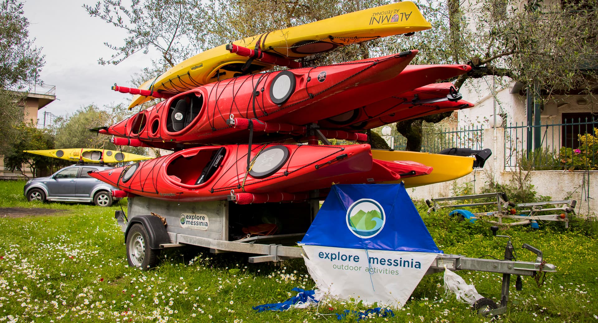Red and yellow sea kayaks stacked on a trailer with an Explore Messinia banner in a flowering garden