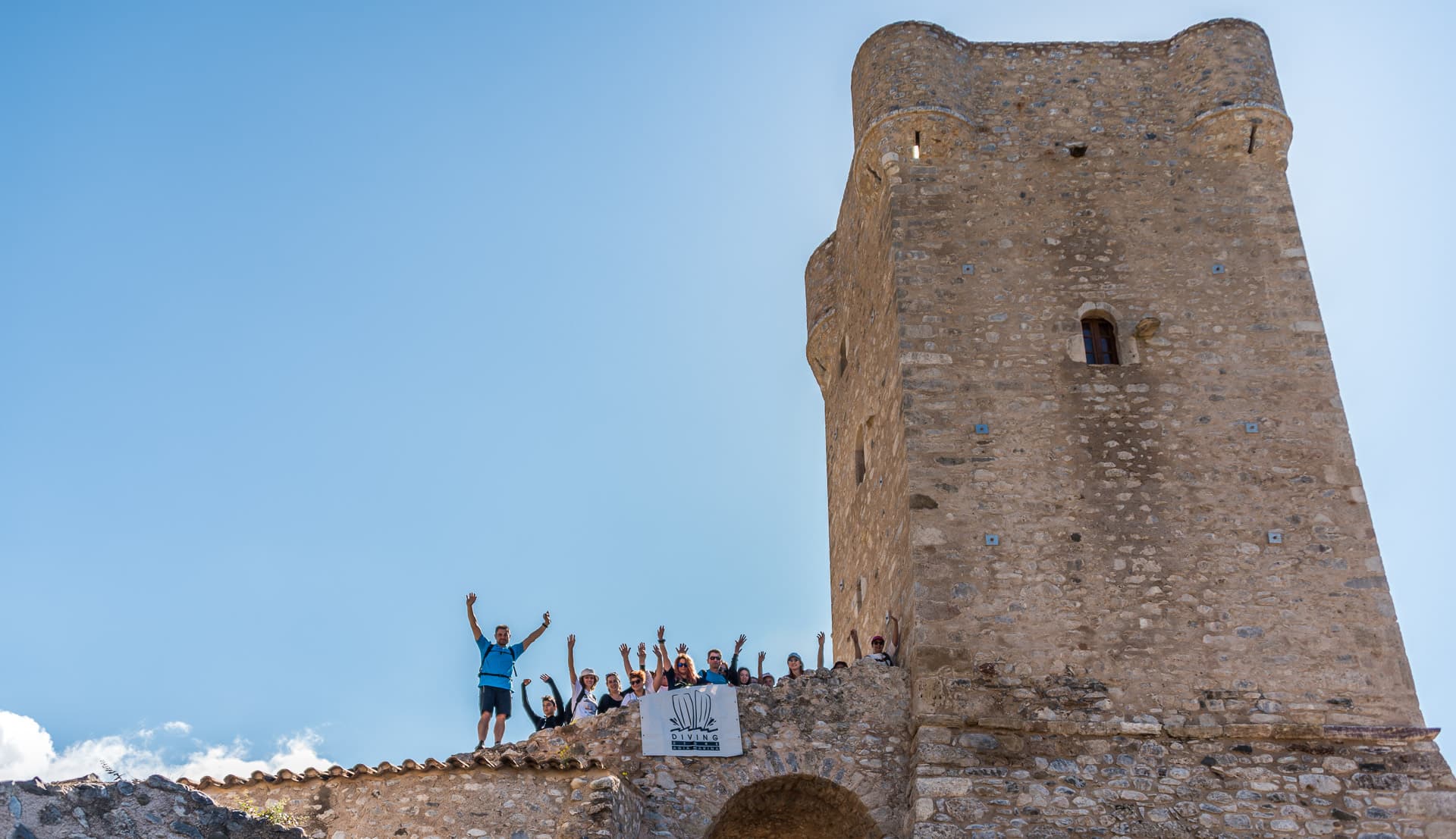 Hikers raising their arms on a stone wall below the tall medieval tower of Kardamyli old town