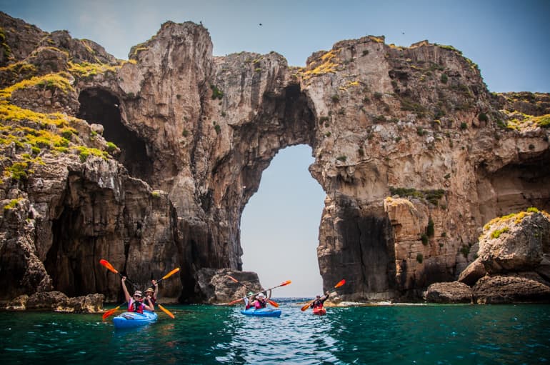 Kayakers paddling toward a dramatic natural sea-rock arch in Navarino bay