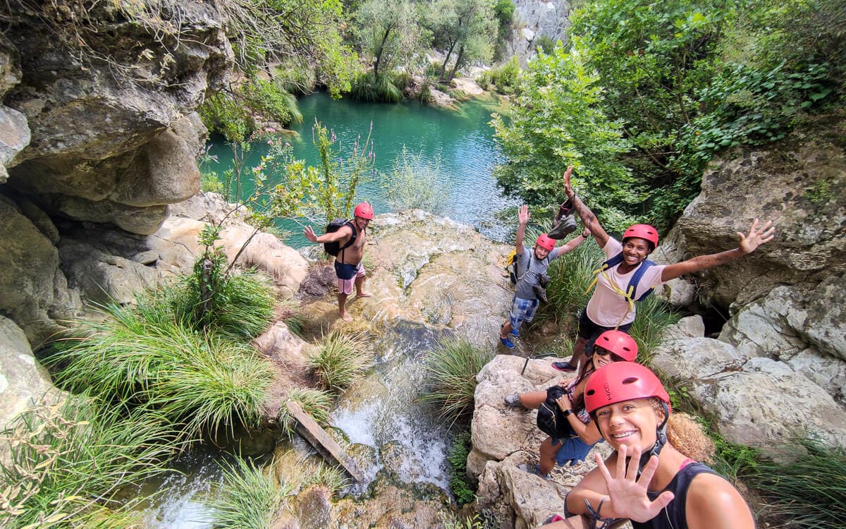 Group of canyoners in red helmets waving from rocks above an emerald pool at Polylimnio