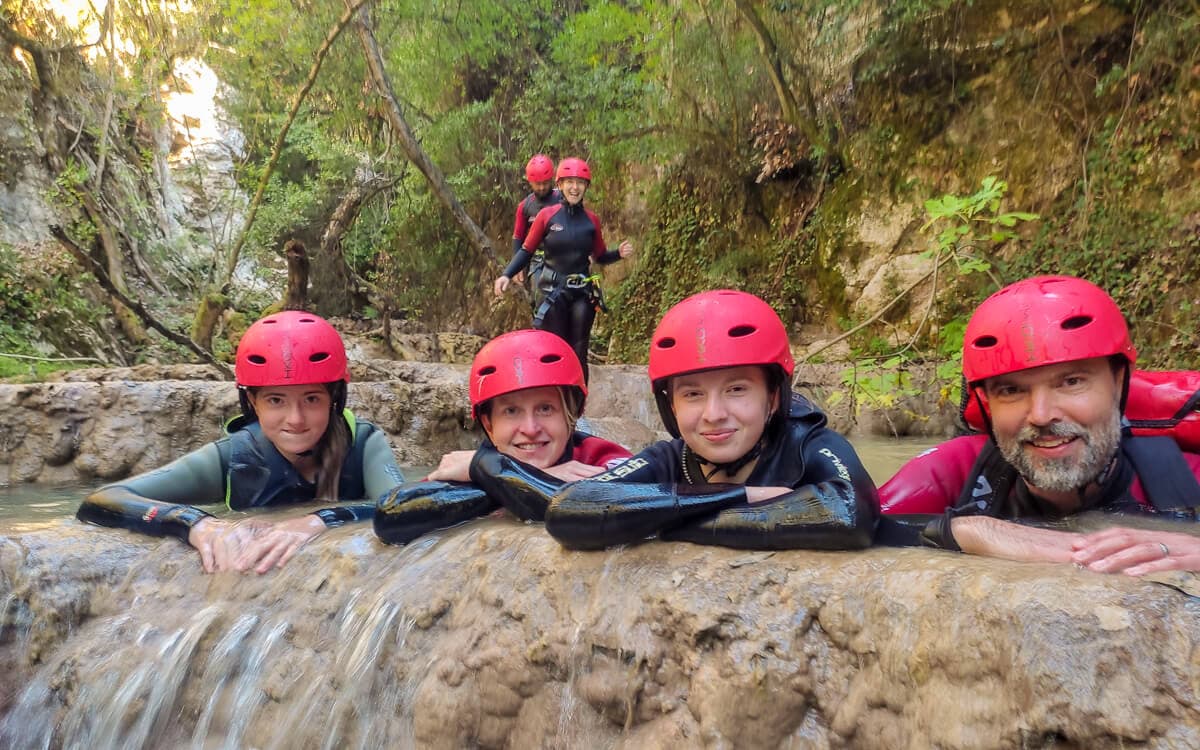 Four canyoners in red helmets and wetsuits resting at the lip of a small waterfall at Polylimnio