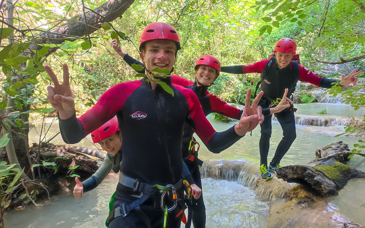 Cheerful canyoners in red helmets flashing peace signs by a forest stream at Polylimnio