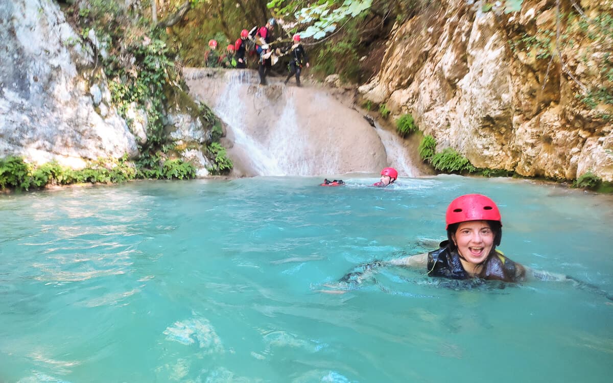 Girl in red helmet swimming in a bright turquoise pool below a waterfall at Polylimnio canyoning