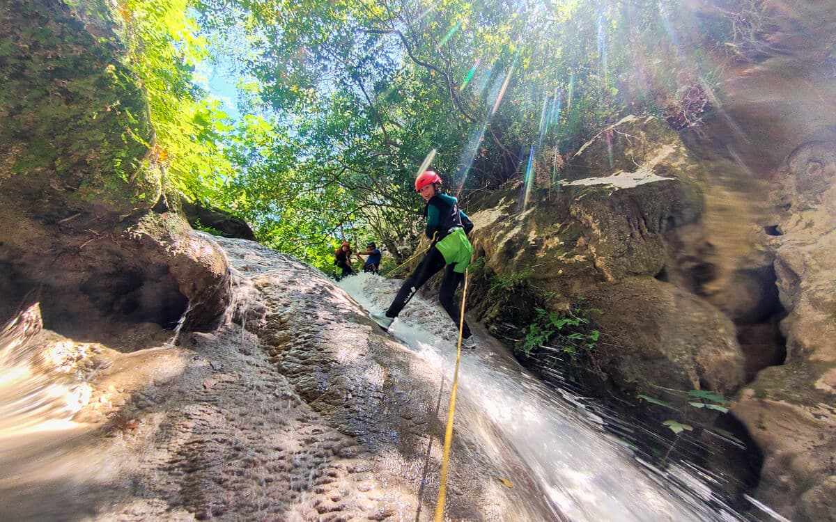 Canyoner in red helmet rappelling down a sunlit rock face on a yellow rope at Polylimnio