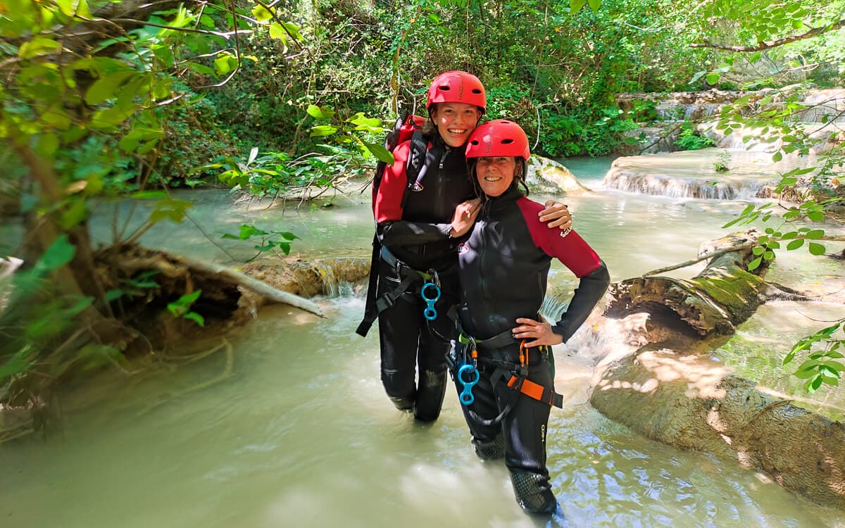 Two women in wetsuits and red helmets smiling in the turquoise pools of Polylimnio canyon