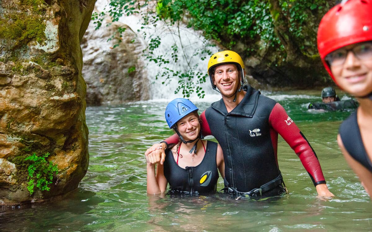 Smiling couple in wetsuits and helmets standing in a green river pool while canyoning at Polylimnio