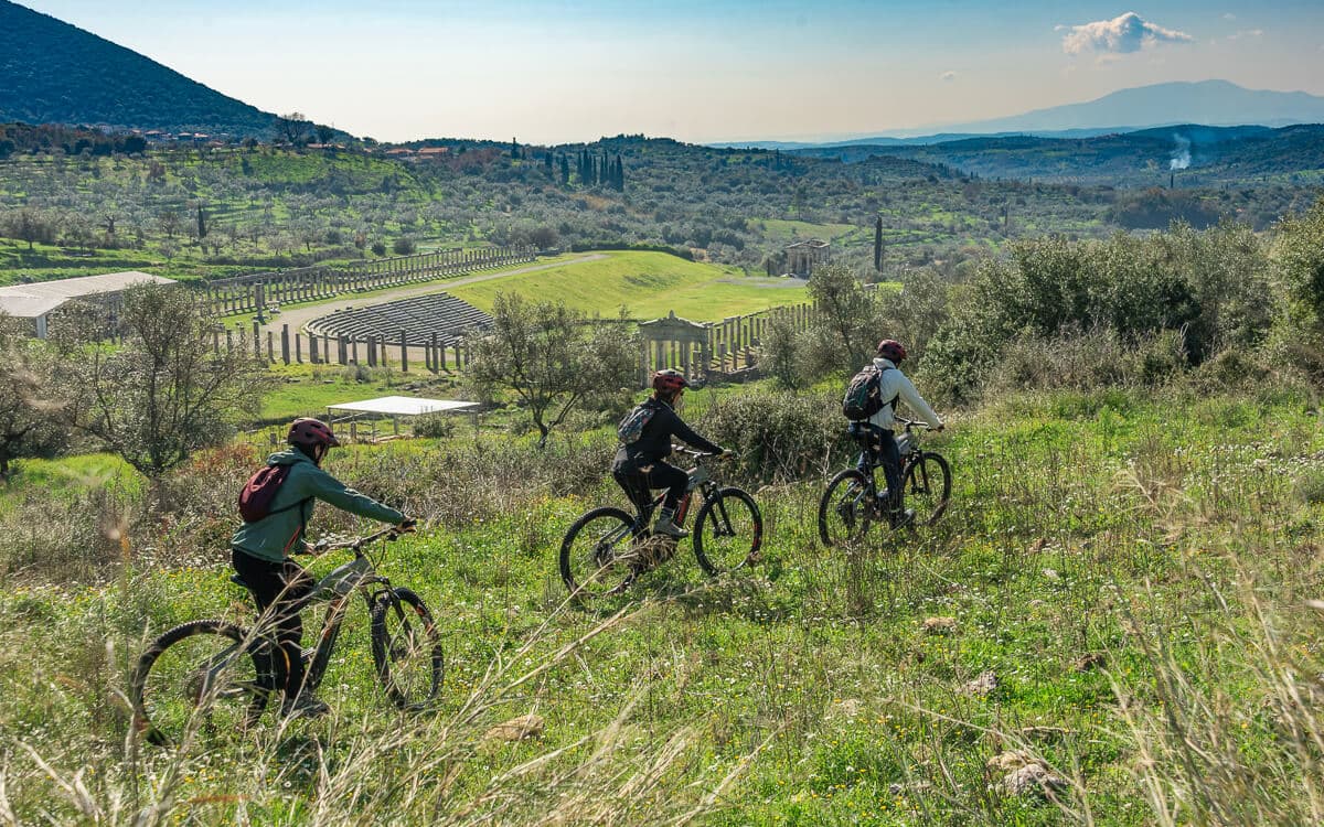 Three e-bikers crossing a hillside meadow above the ancient stadium ruins of Ancient Messene