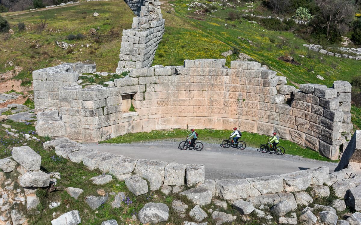 Three e-bike riders pass the curved ancient stone walls of Ancient Messene's stadium ruins