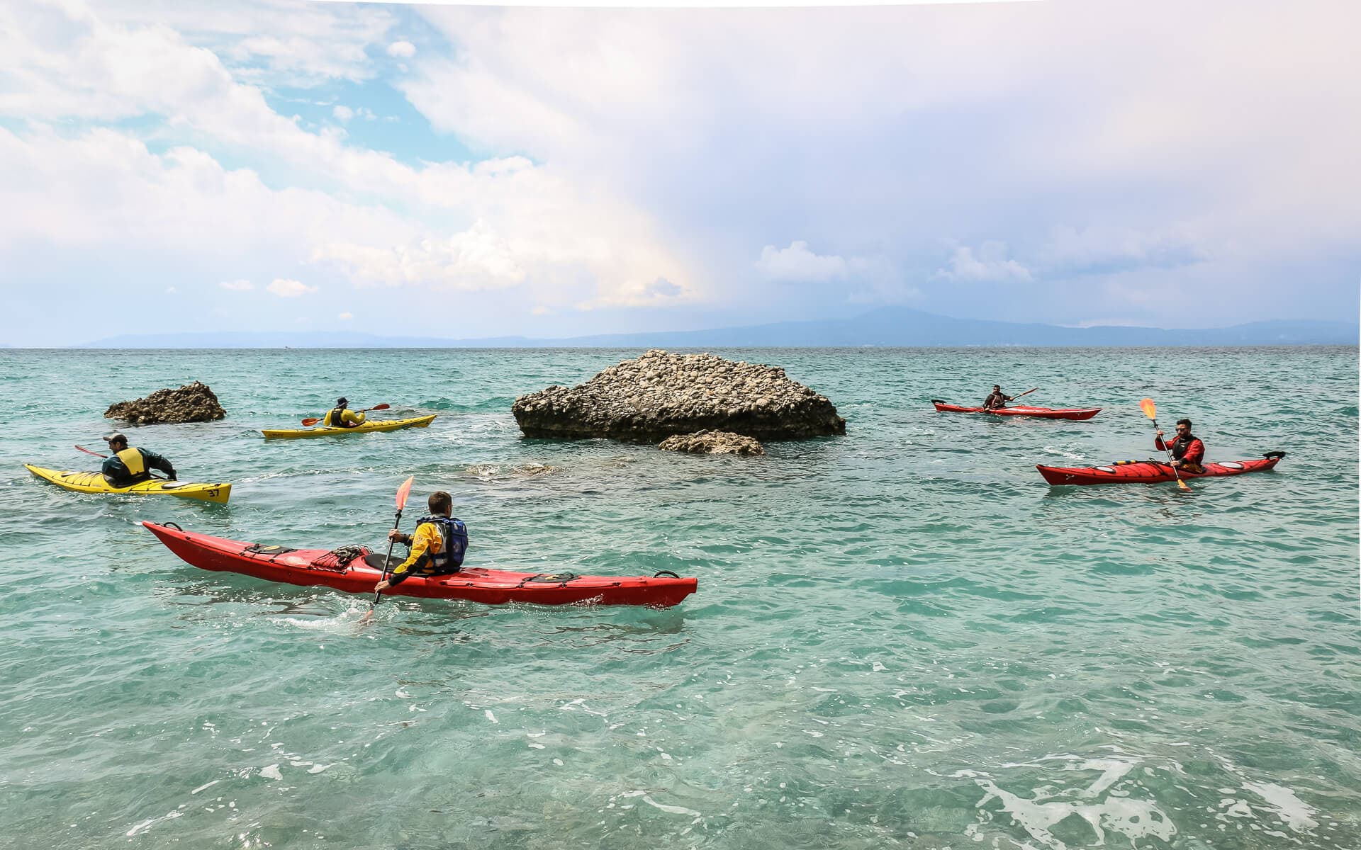 A group of sea kayakers navigating through clear turquoise water near rocky outcroppings.