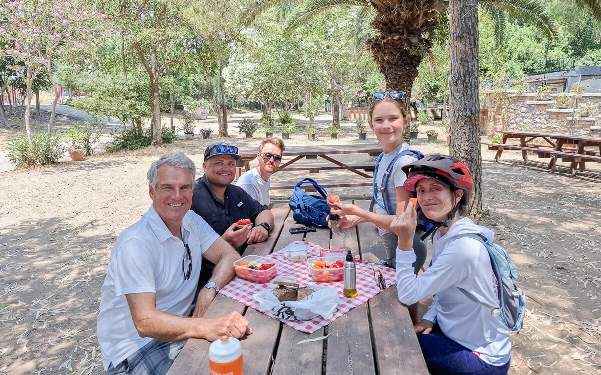 Cycling group sharing snacks at a wooden picnic table under trees near Kalamata