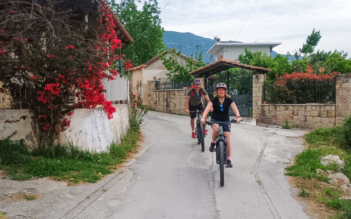 Two cyclists riding down a village lane past a stone wall draped in red bougainvillea near Kalamata