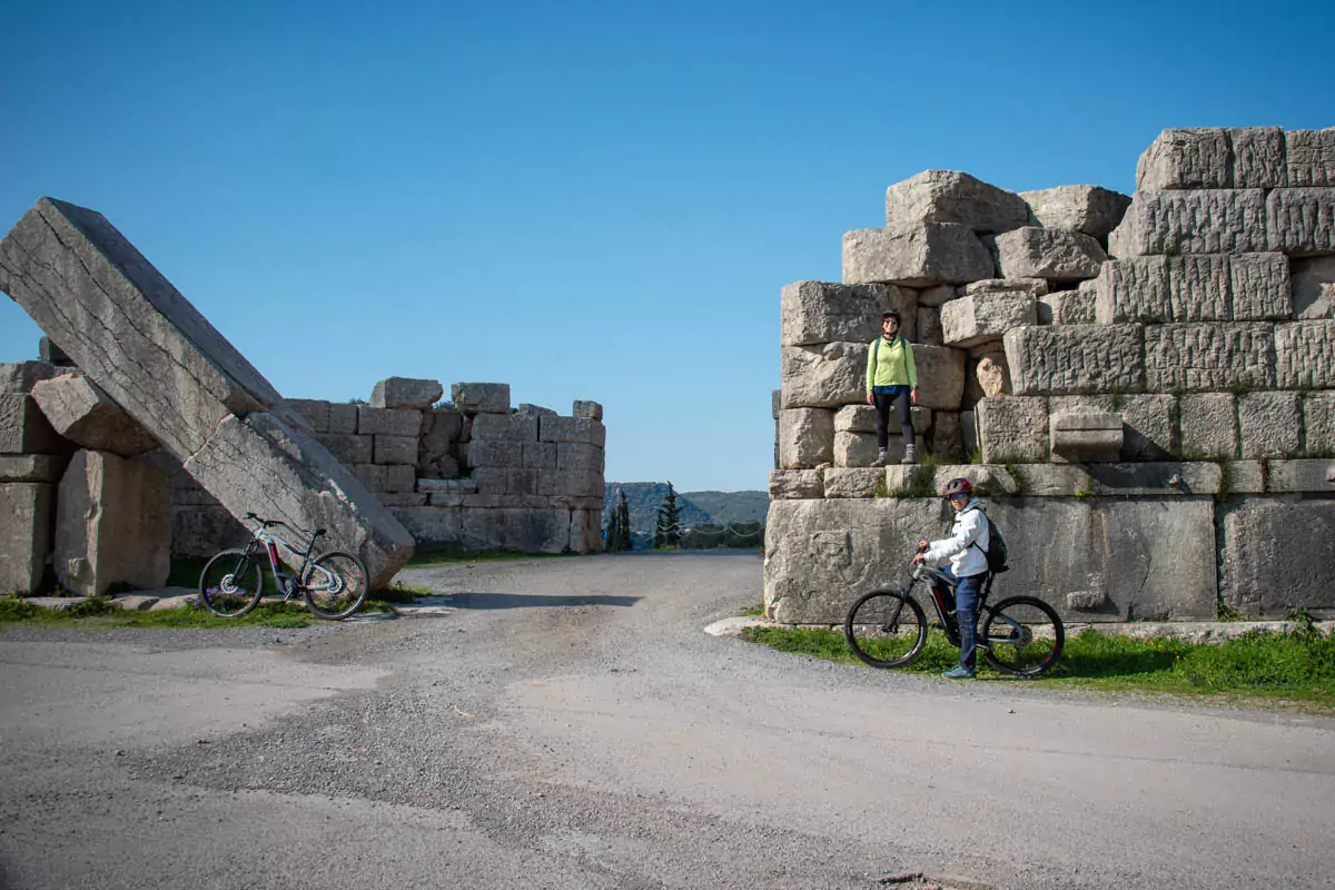 Explore Messinia-A captivating image of ancient Messene featuring a couple standing beside their e-bikes under a historic stone gate. The background showcases the iconic ruins of the ancient site, surrounded by verdant hills and clear blue skies. This scene perfectly embodies the allure of E-bike tours in Greece, blending historical exploration with scenic beauty and adventure.