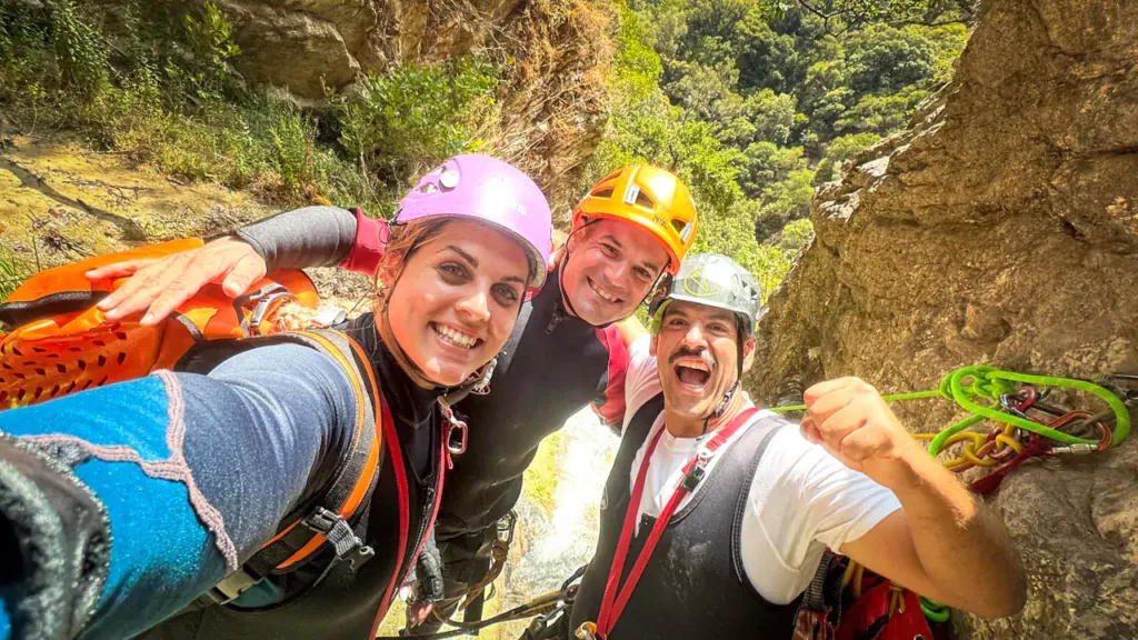 Three smiling canyoning adventurers in helmets and harnesses pose for a selfie in a green gorge