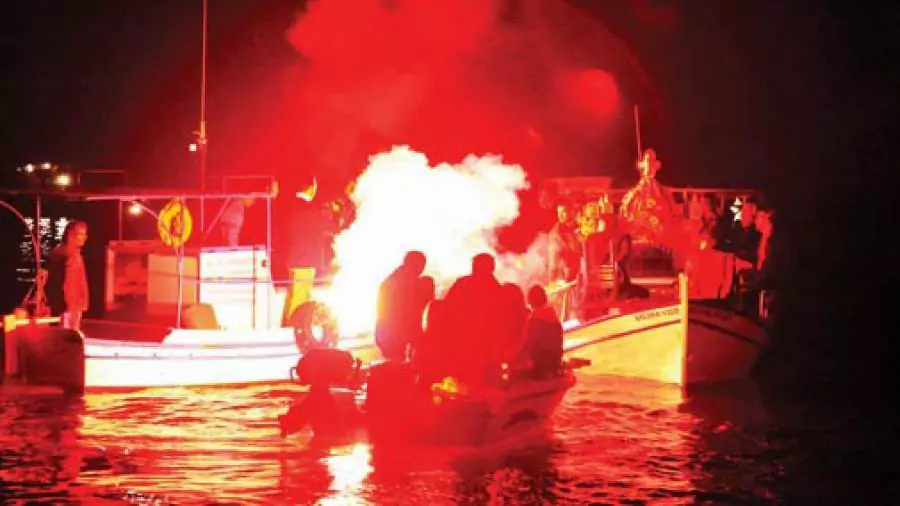 Explore Messinia-A flower-covered epitaph is placed on a fishing boat in a night ceremony, surrounded by smaller boats lit with candles in Kalamata's port.