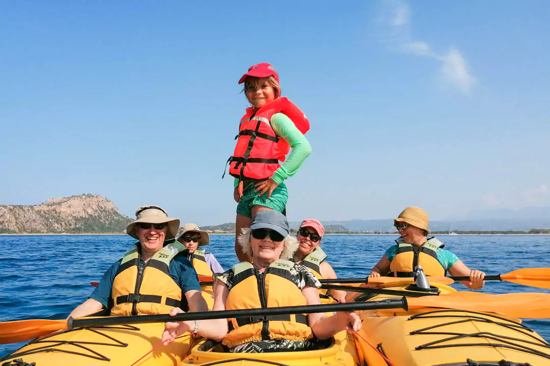 Explore Messinia-A young child with a life jacket, standing on kayaks, exploring a peaceful bay in Messinia, Greece, emphasizing kid-friendly kayaking adventures.