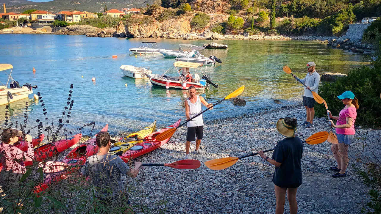 A kayaking guide demonstrating safety instructions and paddling techniques to a group of attentive participants, with kayaks and calm waters in the background.