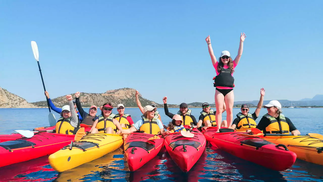 Explore Messinia-A smiling family in life jackets paddling together in a tandem kayak on calm turquoise waters, with scenic cliffs in the background, showcasing the joy of family kayaking.