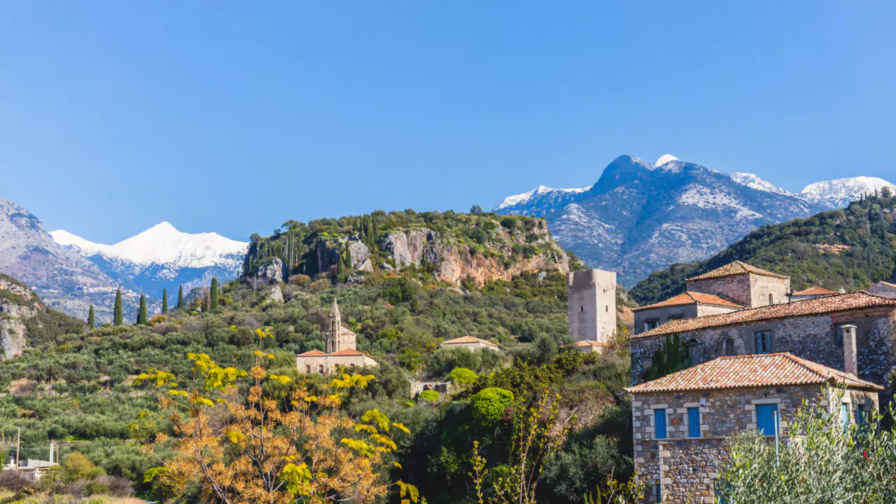 Picturesque villages framed by the majestic Taygetos Mountains. Snow-capped Taygetos Mountains behind green hills, traditional stone buildings with red-tiled roofs, and cypress trees in Mani Greece.